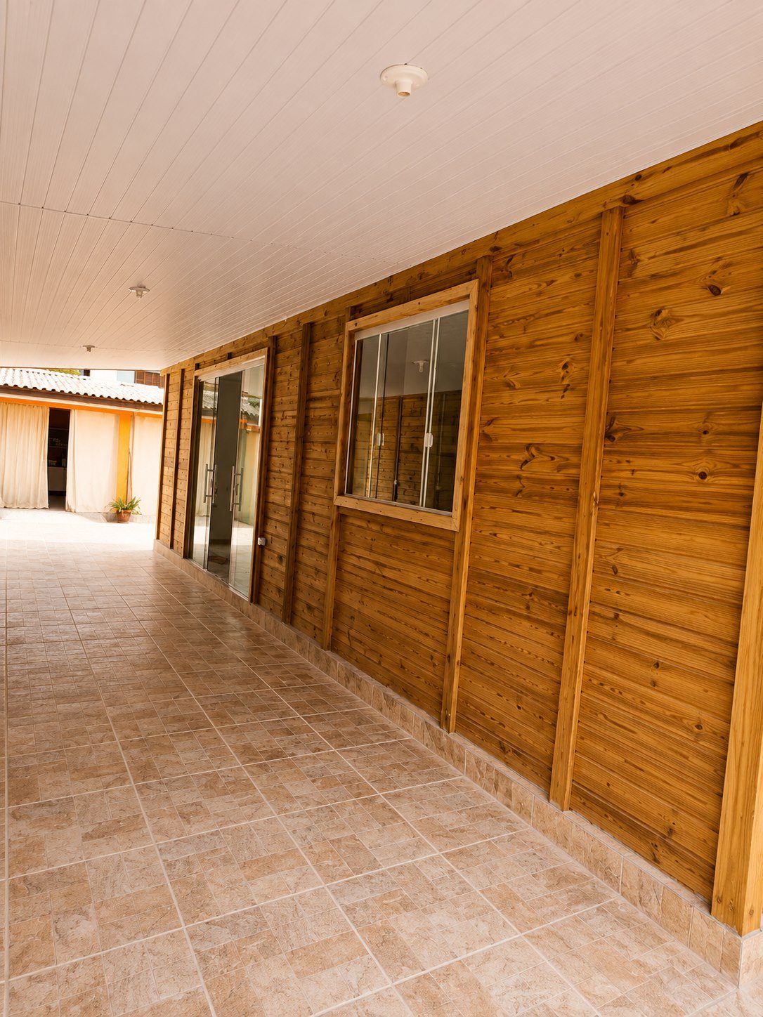 Corredor coberto com parede de madeira, janela de vidro e piso ceramico